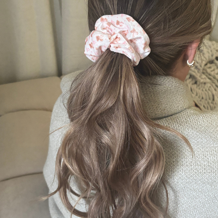 Woman with wavy brown hair in a ponytail wears a beige, fluffy teddy scrunchie while seated on a neutral couch.