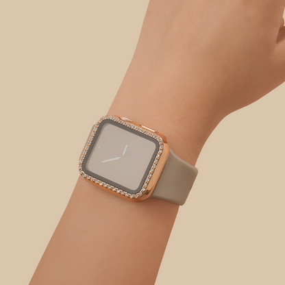 A close-up of a wrist wearing the Coconut Lane 2-in-1 Apple Watch Case & Built-In Screen Protector - Rose Gold Diamante, featuring a rectangular face. The watch case has a rose gold diamante frame adorned with small, sparkling stones around the edges and is paired with a light gray strap. The background is a solid beige color.