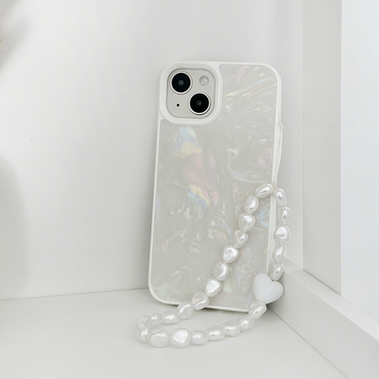 A white smartphone with a shimmering, iridescent case is propped upright against a white corner. The phone case boasts Coconut Lane's stylish Beaded Phone Strap - Chunky Pearls, adorned with irregularly shaped white pearls and ending in a small white heart. The background and surface are also white, creating a minimalist aesthetic.