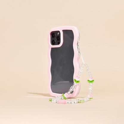 A smartphone case with a wavy pink edge, protecting a phone with triple rear cameras, is shown standing upright. Attached to the case is the Beaded Phone Strap - Cute Tulips by Coconut Lane, featuring small green leaves and pink beads that resemble cherries. This charming phone accessory stands against a plain beige background.