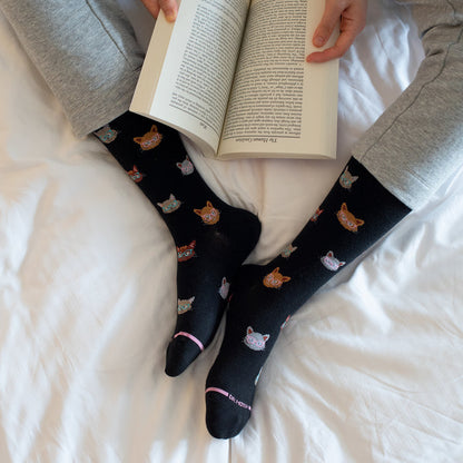 Knee-high black compression socks with colorful cat faces and pink accents, worn while reading a book.