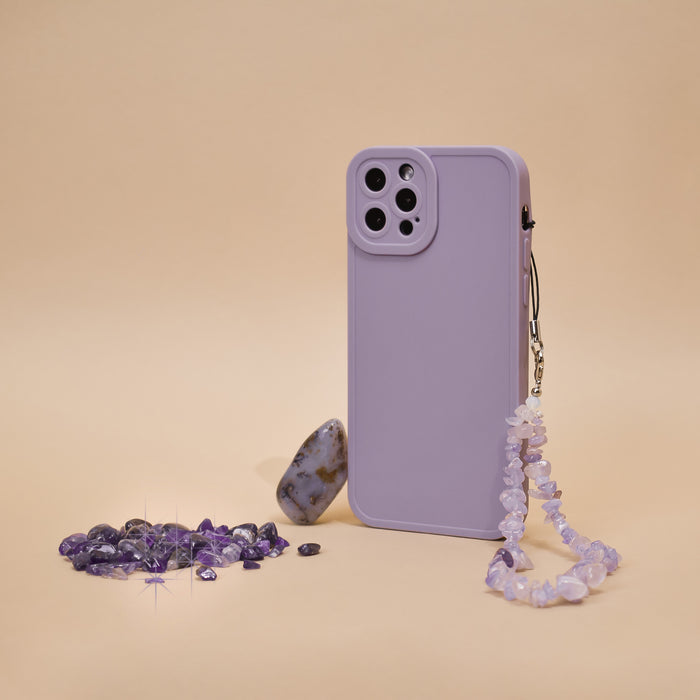 A phone case in a vibrant lilac shade with Coconut Lane's Crystal Phone Strap adorned with small beads stands upright against a beige background. Scattered around the stylish accessory are amethyst crystals and one larger, smooth stone with earthy tones.