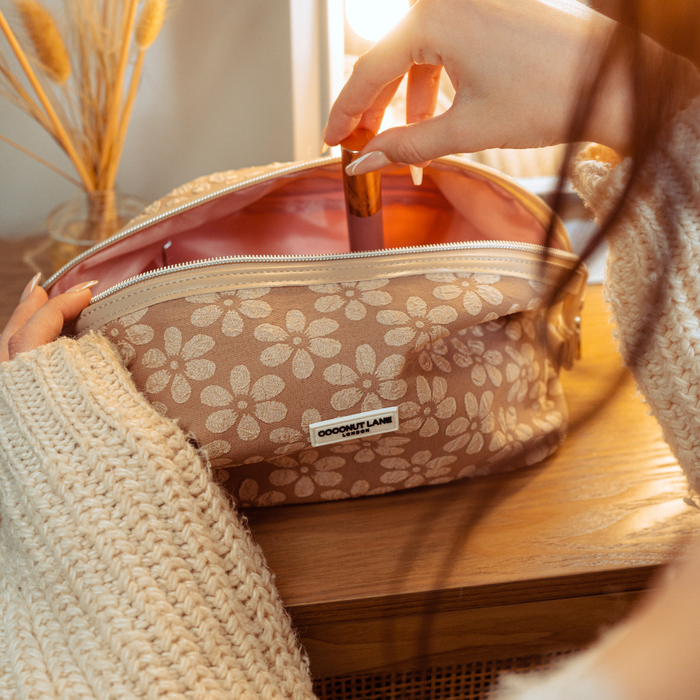 Lipstick being placed into a mocha floral Coconut Lane expandable makeup bag on wooden surface near dried flowers.