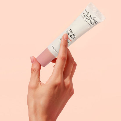 Hand holding a tube of 'The Jojoba Company' Firming Eye Balm against a pink background.
