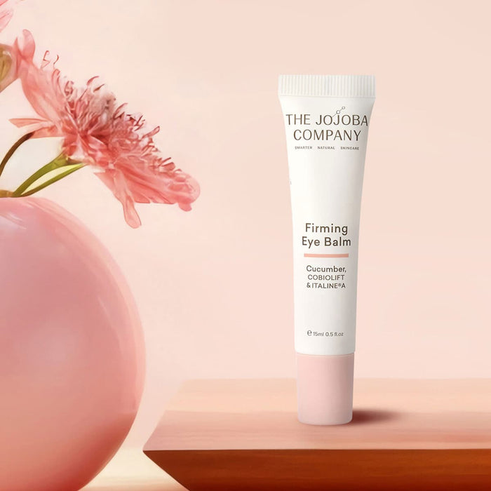 Firming eye balm tube from The Jojoba Company with a pink flower on a peach background.