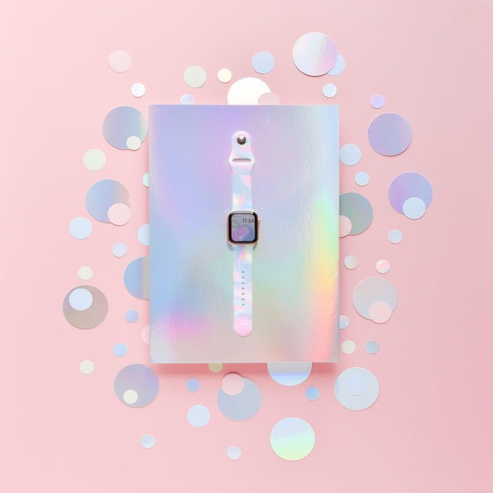 A white smartwatch with a square screen is centered on a holographic, iridescent background. The Holo Bubble Apple Watch Strap by Coconut Lane stands out, complementing the backdrop's mix of circular cutouts in various sizes. The entire scene is enveloped in a soft, pastel pink tone.