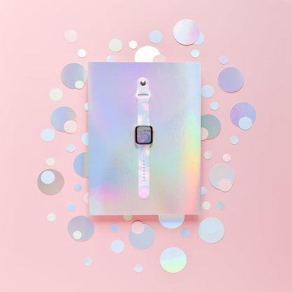 A white smartwatch with a square screen is centered on a holographic, iridescent background. The Holo Bubble Apple Watch Strap by Coconut Lane stands out, complementing the backdrop's mix of circular cutouts in various sizes. The entire scene is enveloped in a soft, pastel pink tone.