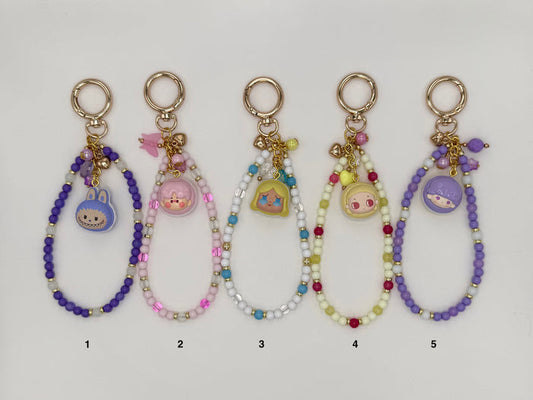 Five numbered kawaii wrist strap keychains on a white background, each with a gold spring ring clasp and a macaron-shaped dome charm containing a chibi character face. Variants include a purple strap with blue bunny charm (1), pink and mauve strap with pink piggy charm (2), white and blue strap with yellow Alice-style charm (3), yellow-green and red strap with pink chibi charm (4), and an all-purple strap with blue character charm (5).