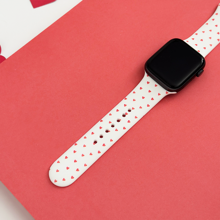 White silicone Apple Watch strap with small heart pattern, displayed on a pink background.