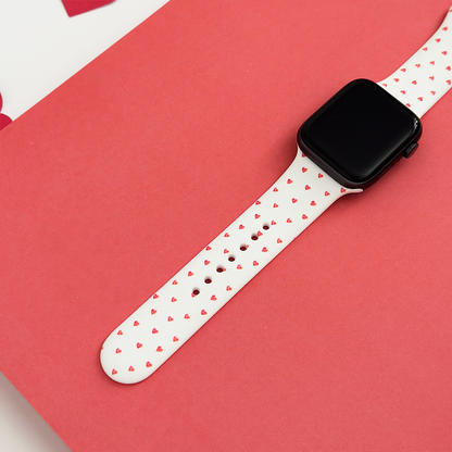 White silicone Apple Watch strap with small heart pattern, displayed on a pink background.