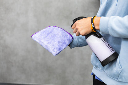 Person holding a white iK spray bottle with a purple minx royale microfiber cloth against a gray background