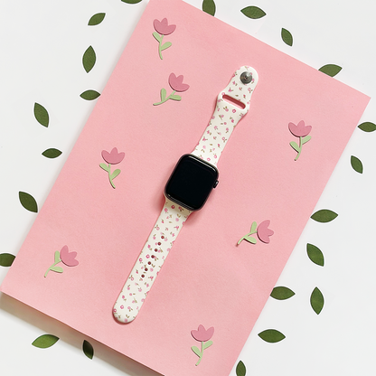 Pink floral Apple Watch strap with watercolor flower pattern, shown on a pink background with green leaves.