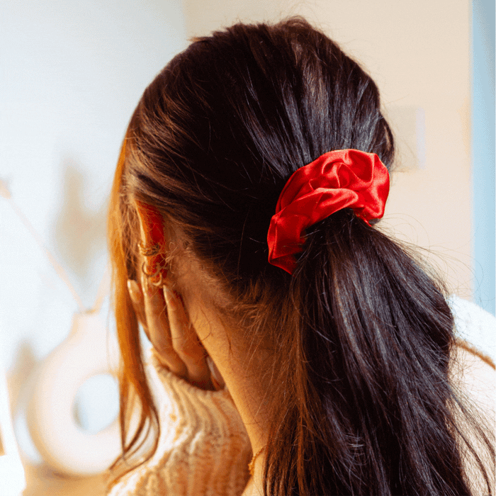 Woman with long brown hair wearing a rose red satin bow scrunchie, resting her hand on her face.
