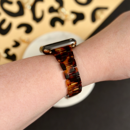 Wrist with Coconut Lane Slim Luxe tortoiseshell Apple Watch strap, thin and glossy, shown against a patterned background.