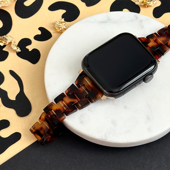 Slim tortoiseshell Apple Watch strap with gold hardware on a white marble dish atop a patterned background.