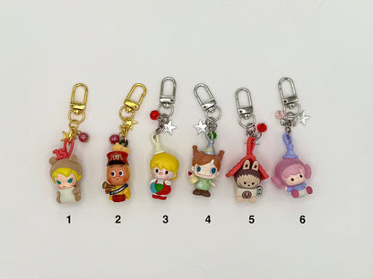 Six numbered character keychains on a white background. Features: (1) blonde girl in a bear hood with gold clasp, bell and star charms, (2) brown character in a red marching band uniform with gold clasp, (3) blonde boy holding a ball with silver clasp and bead charms, (4) freckled girl in brown outfit with silver clasp, (5) hedgehog in a red house hat with clock detail, (6) pink boy in lavender outfit with crystal charms.