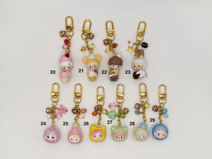 Ten numbered kawaii character keychains with gold clasps on a white background. Includes a pink elf girl (20), cream mouse in ice cream cone costume (21), mushroom-cap boy (22), white wolf with star charms (23), pink ghost figure (24), pink potion bottle (25), yellow bear face (26), green hooded character (27), and two clear dome figures with characters inside (28–29), all with decorative bead and chain accents.