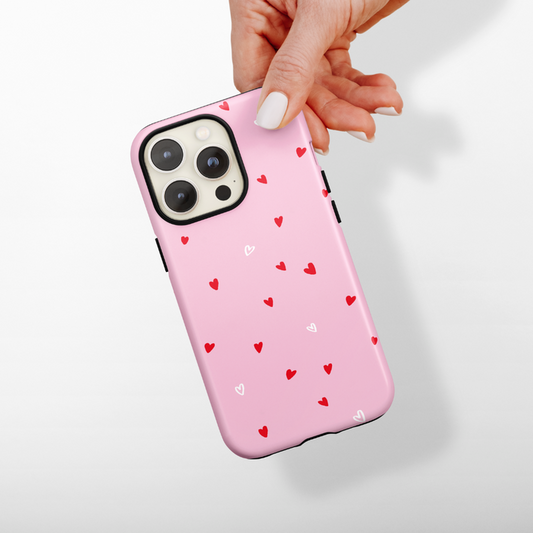 A hand with white nail polish is holding an iPhone adorned with Coconut Lane's Tough Phone Case - Cute Hearts, featuring small red and white hearts against a pink backdrop. The setting is a plain white background.