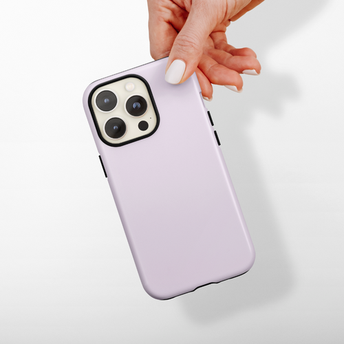 A hand with manicured nails is holding a smartphone with a light lilac "Tough Phone Case - NAKD Lilac" by Coconut Lane. The phone has a triple camera setup on the back. The background is plain and light-colored, casting a shadow of the hand and phone.
