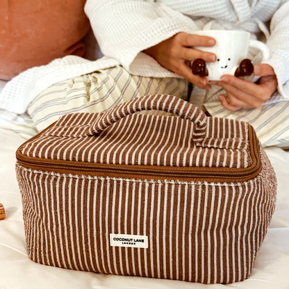 Chestnut striped vanity case by Coconut Lane on a bed, with pajama-clad person holding a mug in the background.